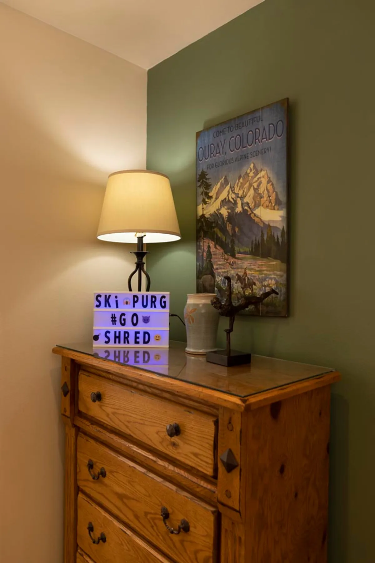A cozy corner featuring a lamp, mountain artwork, and a dresser topped with a ceramic vase and decorative branch. The setup exudes a welcoming, personalized vibe, inviting guests to unwind and create their own sanctuary after a productive day.
