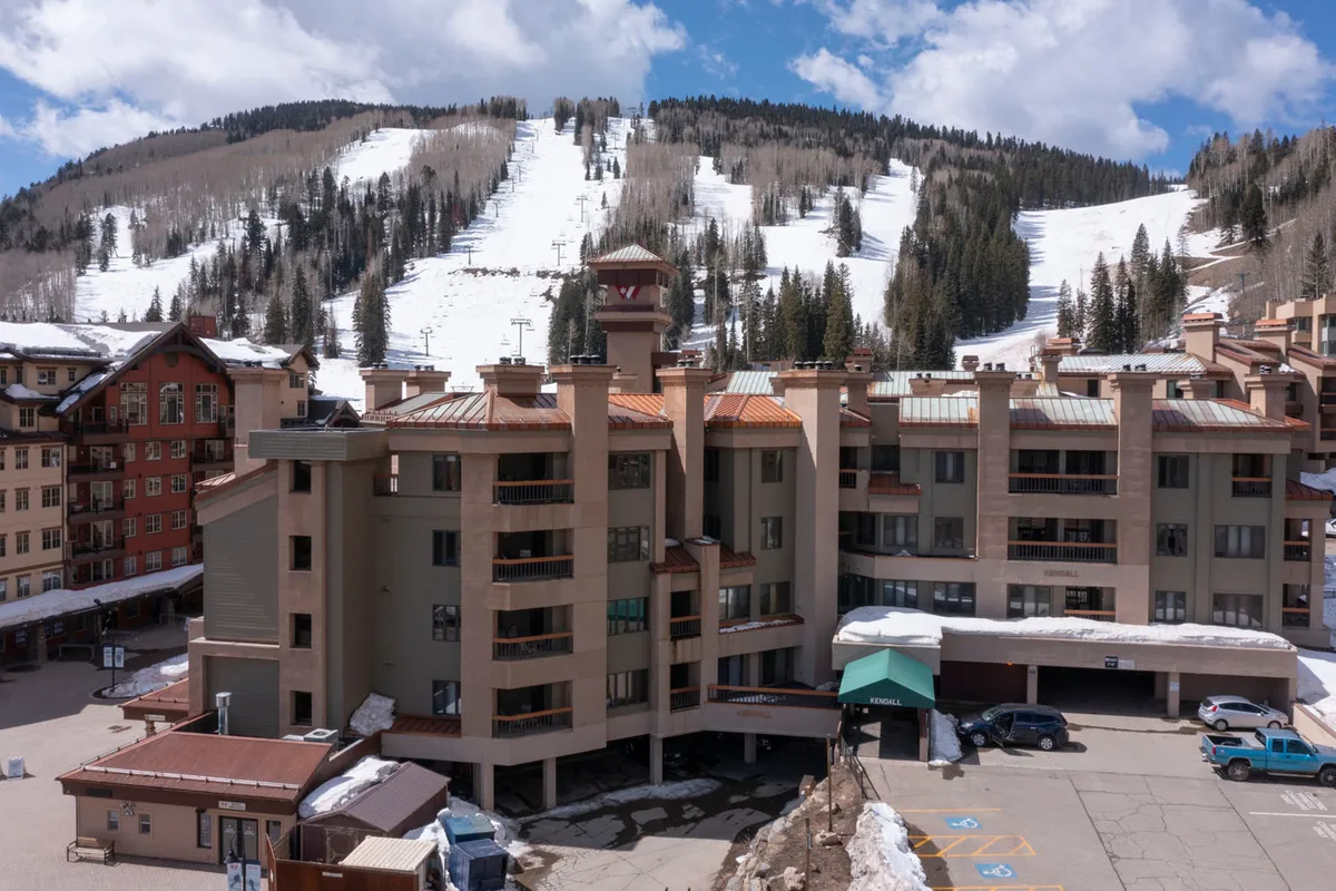 This exterior image showcases the building's architectural details with a visible parking area in the foreground, set against a striking backdrop of snowy mountains. The scene is inviting, hinting at easy access and the comfort of arriving at a resort with available parking and beautiful surroundings.