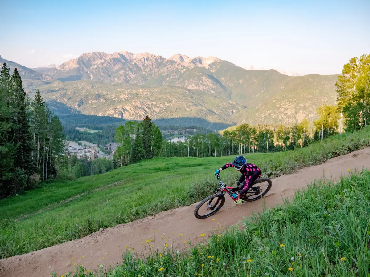 A mountain biker rides along a winding dirt trail, surrounded by lush greenery and towering mountains under a clear sky. The scene radiates energy and adventure, capturing the essence of outdoor activities like biking and hiking in a stunning natural setting.
