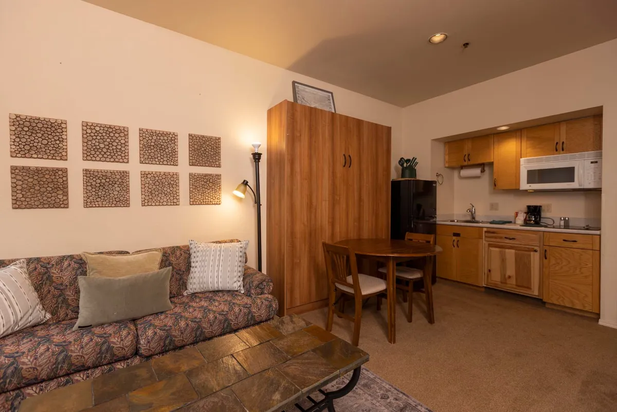 This image showcases a welcoming studio interior with a comfortable sofa, a dining table, and a fully equipped kitchen area visible in the background. Thoughtful wall art and decor create a soothing, home-like atmosphere, and the layout highlights both the living and dining zones, perfect for work or relaxation.