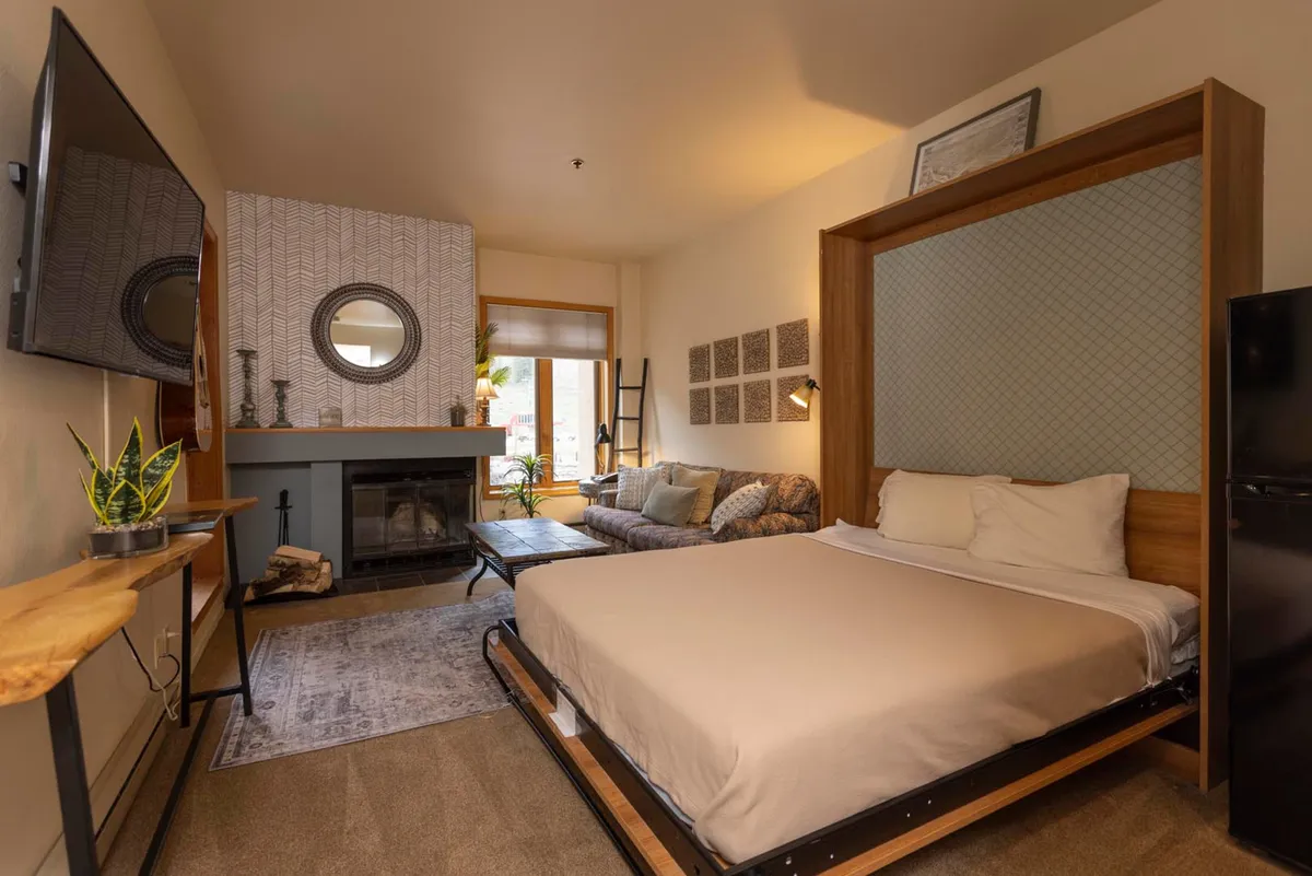 A spacious yet intimate area where a bed, sofa, and wood-burning fireplace coexist, framed by decorative details and a TV. Soft, natural light filters through large windows, fostering a comfortable, welcoming environment ideal for unwinding and spending quiet moments together.