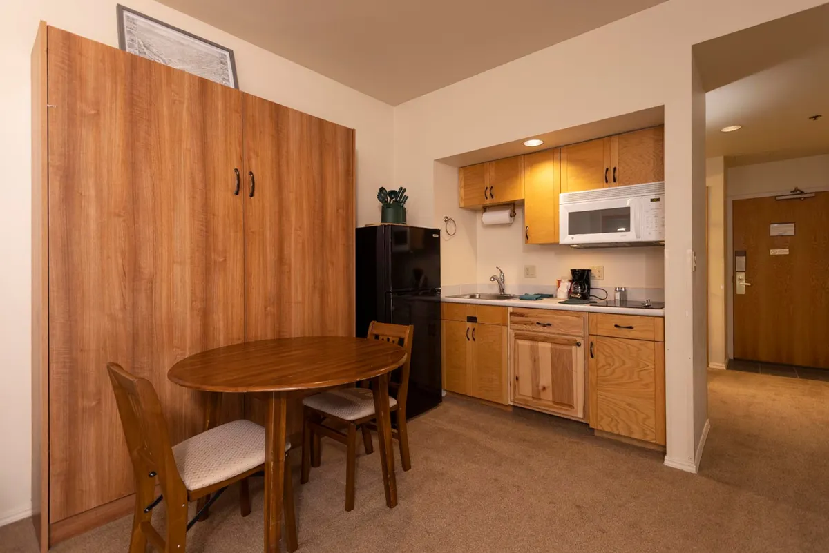 This image showcases a welcoming kitchen and dining nook, featuring a wooden dining table, a compact kitchenette with essential appliances, a folding cabinet, and a black refrigerator. The arrangement encourages intimate gatherings and casual meals, with natural light highlighting the space’s cozy, functional design.