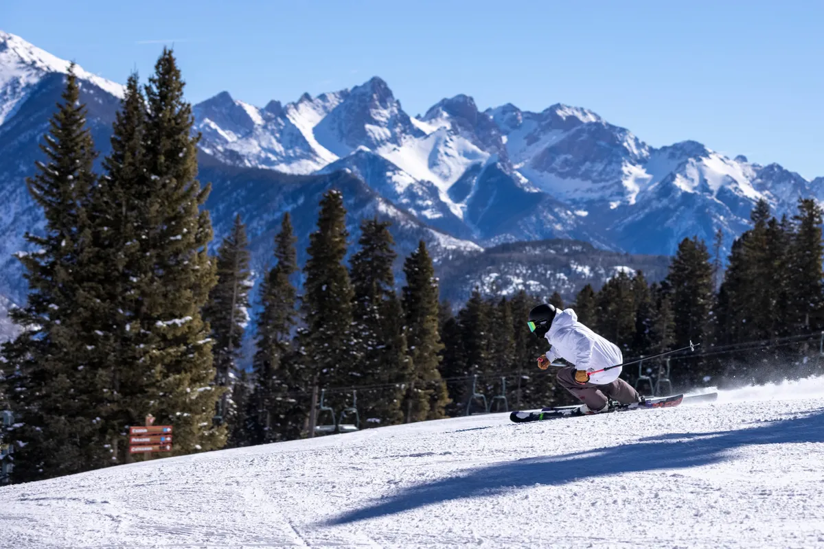 A skier carves through fresh powder down a snow-covered mountainside, framed by majestic evergreens and distant peaks. The crisp winter setting highlights the thrill of skiing and snowboarding at a renowned mountain resort.