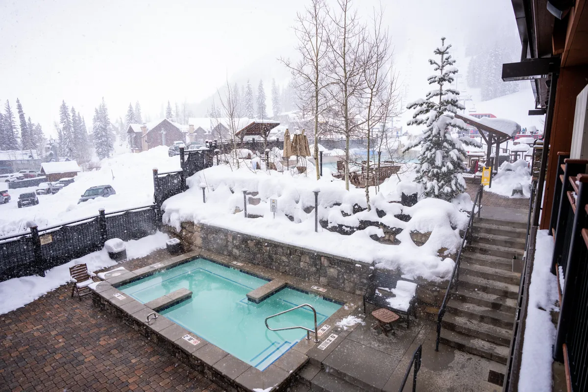 An exterior shot featuring a steaming hot tub surrounded by a snowy landscape, flanked by trees and accessed via stone steps. The ambiance is tranquil and serene, inviting guests to soak under the stars or enjoy the calm of winter after a day of adventure.