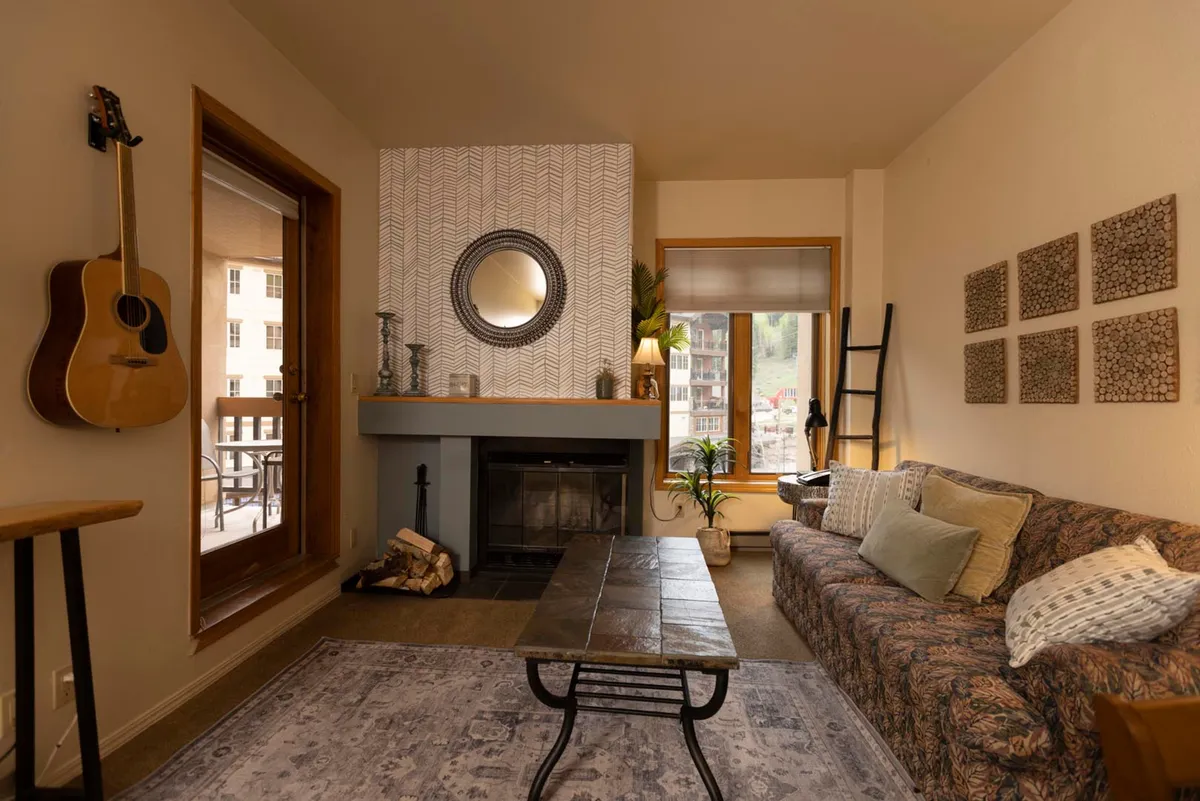 The image shows a cozy living space in the condo, centered around a glowing wood-burning fireplace. A comfortable sofa sits nearby, with a guitar resting against it, hinting at laid-back music sessions. A coffee table holds a book and mug, and a large mirror reflects warm sunset light streaming through the window, creating an inviting and peaceful ambiance.