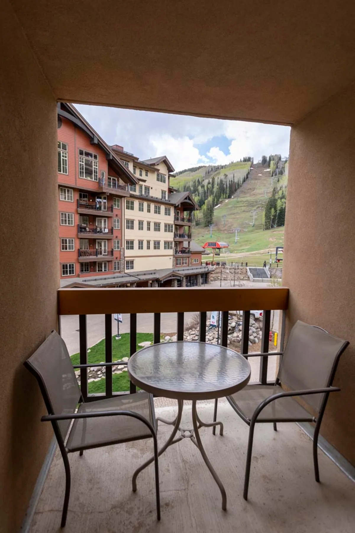 An exterior shot of a private balcony featuring two chairs and a table, overlooking the ski resort with panoramic mountain views. The scene embodies resort relaxation and immediate access to the outdoors, suggesting proximity to amenities like the Village Plaza.