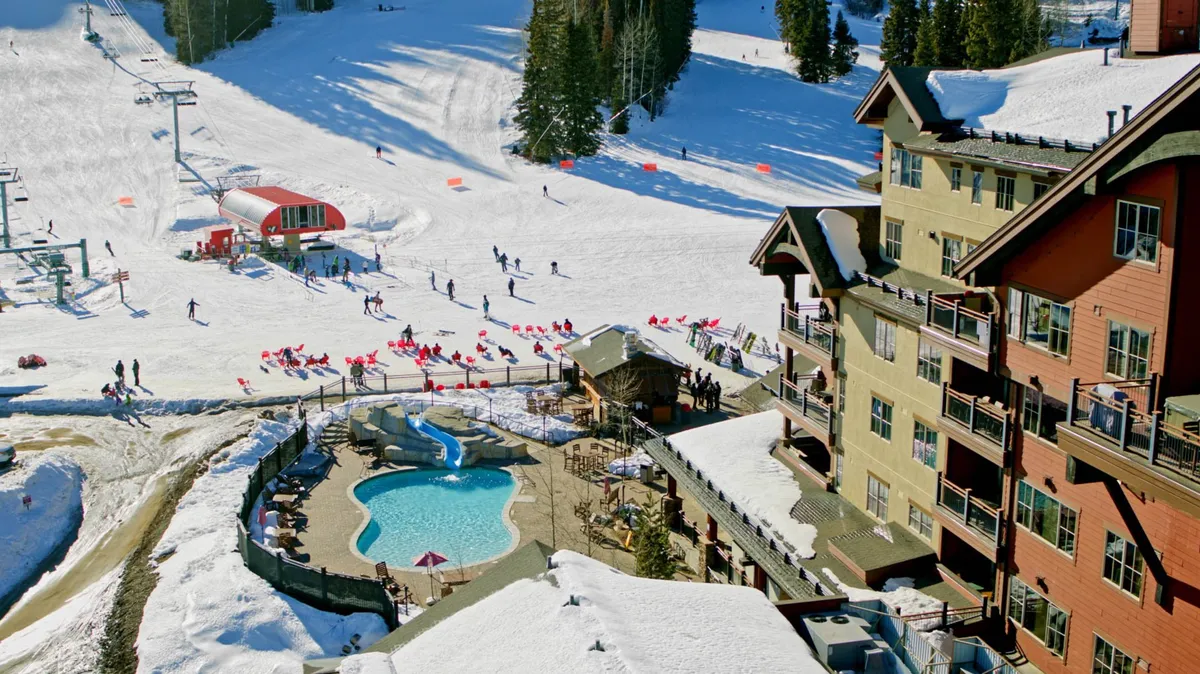 A wide exterior shot showing a lively swimming pool adjacent to ski slopes, with the lodge building and ski lifts in the background. The area is vibrant, blending the fun of swimming with the excitement of mountain activities.