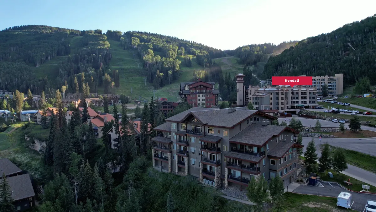This image captures a sweeping mountain backdrop with residential buildings nestled among trees, and a parking area visible in the foreground. The atmospheric setting hints at an inviting mountain community surrounded by natural beauty, making it ideal for both adventure and relaxation.