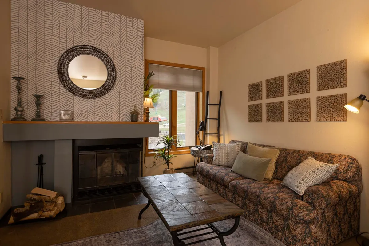 A cozy living area featuring a wood-burning fireplace as the central focus. Plush sofas are arranged invitingly around a coffee table, with decorative wall art and a round mirror enhancing the intimate atmosphere. Large windows provide glimpses of the alpine surroundings, while the fire casts a warm, relaxing glow.